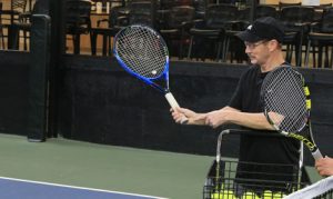 An adult tennis player practices his swing on an indoor court, focusing on improving his technique during a training session. He prepares for an adult tennis league, with a basket of tennis balls by his side for repetitive drills.