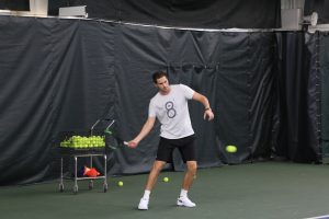 A male tennis player trains for an adult tennis league indoors, hitting a tennis ball with precise focus. Behind him, a cart full of tennis balls is ready for use, supporting the player's efforts to sharpen his game.