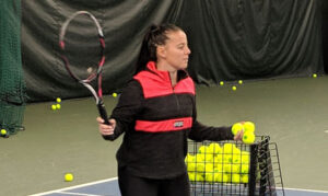 A female tennis player holds tennis balls in one hand while preparing to hit with the other, during an adult tennis league training session. She is practicing indoors, surrounded by tennis balls and a ball cart for drills.